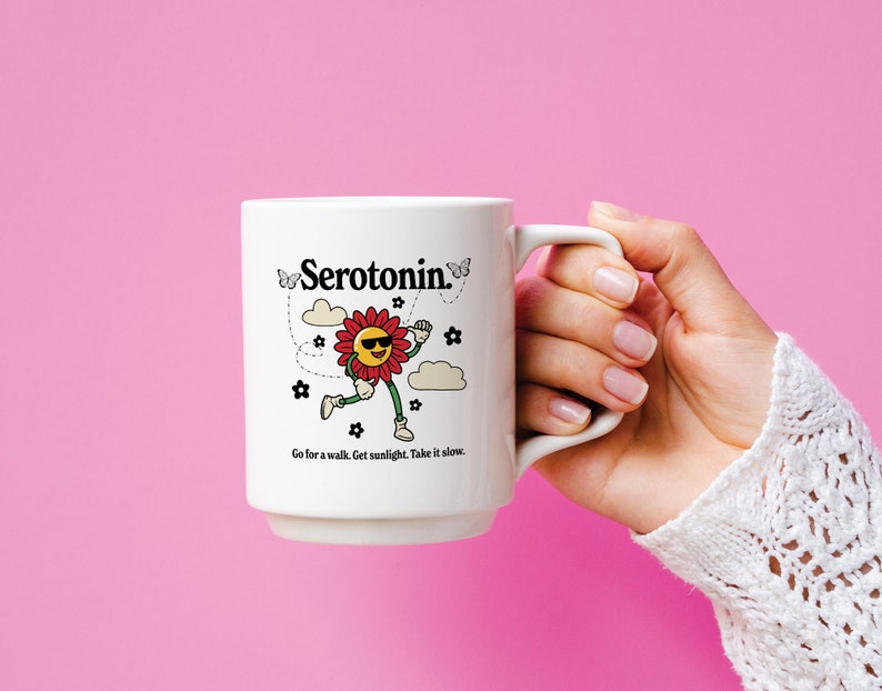 Może przedstawiać: Biały kubek ceramiczny z czarno-białym rysunkiem uśmiechniętego kwiatu w okularach przeciwsłonecznych i napisem "Serotonina" czarnym kolorem. Kubek ma napis "Go for a walk. Get sunlight. Take it slow." nadrukowany na spodzie.