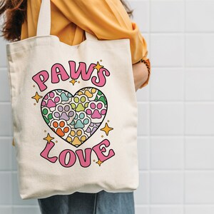 May include: A white canvas tote bag with a pink heart design featuring paw prints in various colors. The text "PAWS LOVE" is printed in pink letters.