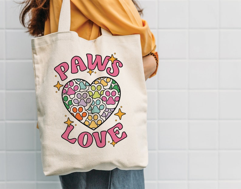 May include: A white canvas tote bag with a pink heart design featuring paw prints in various colors. The text "PAWS LOVE" is printed in pink letters.