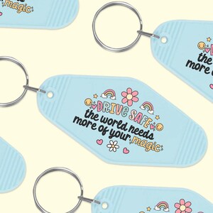 May include: Blue motel key chain with a white and pink design. The keychain features a rainbow, flowers, and the text "DRIVE SAFE the world needs more of your magic".
