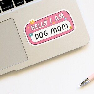 Peut inclure: Autocollant rose avec le texte "Hello I am Dog Mom" en lettres blanches. L'autocollant a une bordure blanche et est d&eacute;cor&eacute; de petites fleurs et d'&eacute;toiles.