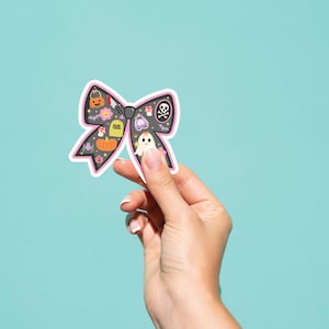 May include: A black bow-shaped sticker with a pink border, featuring Halloween-themed illustrations. The sticker includes a pumpkin, ghost, skull, and tombstone. Held against a light blue background.