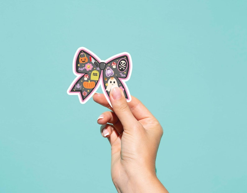 May include: A black bow-shaped sticker with a pink border, featuring Halloween-themed illustrations. The sticker includes a pumpkin, ghost, skull, and tombstone. Held against a light blue background.