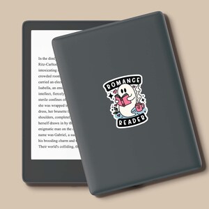 Op de afbeelding: Een zwarte en grijze e-reader met een sticker van een geest die een boek leest en een kopje koffie vasthoudt. De sticker zegt "Romance Reader".