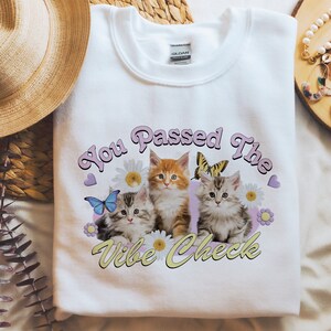 Op de afbeelding: Witte trui met een afbeelding van drie kittens en bloemen. De tekst "You Passed The Vibe Check" is gedrukt in roze en gele letters.