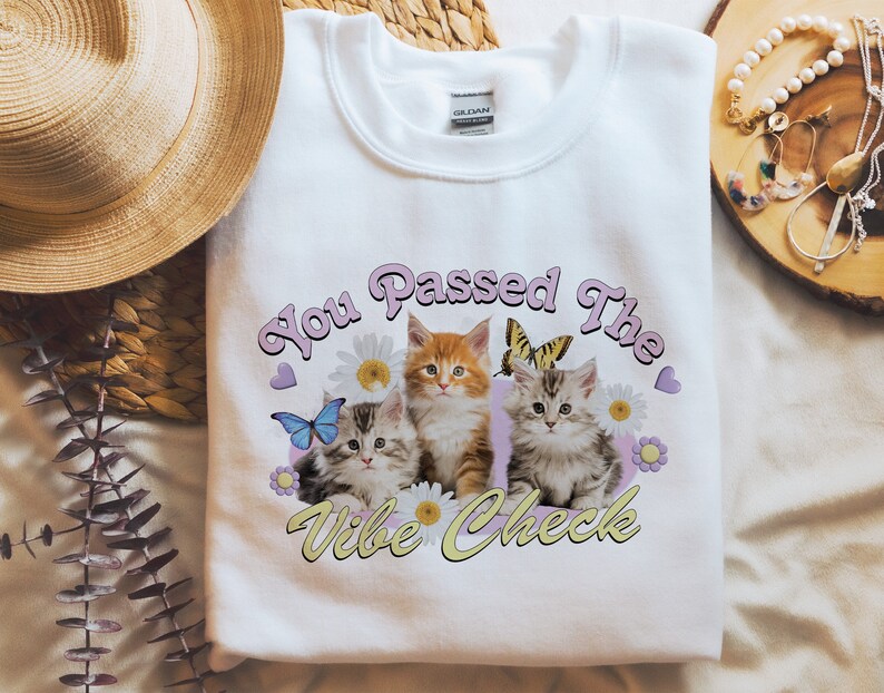 Op de afbeelding: Witte trui met een afbeelding van drie kittens en bloemen. De tekst "You Passed The Vibe Check" is gedrukt in roze en gele letters.