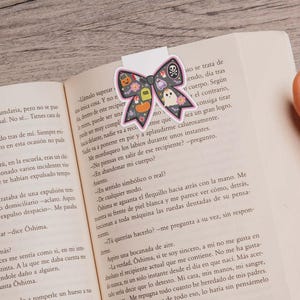 May include: A Halloween bow-shaped bookmark featuring a skull, ghost, pumpkin, and other icons. The bookmark is on an open book with text.