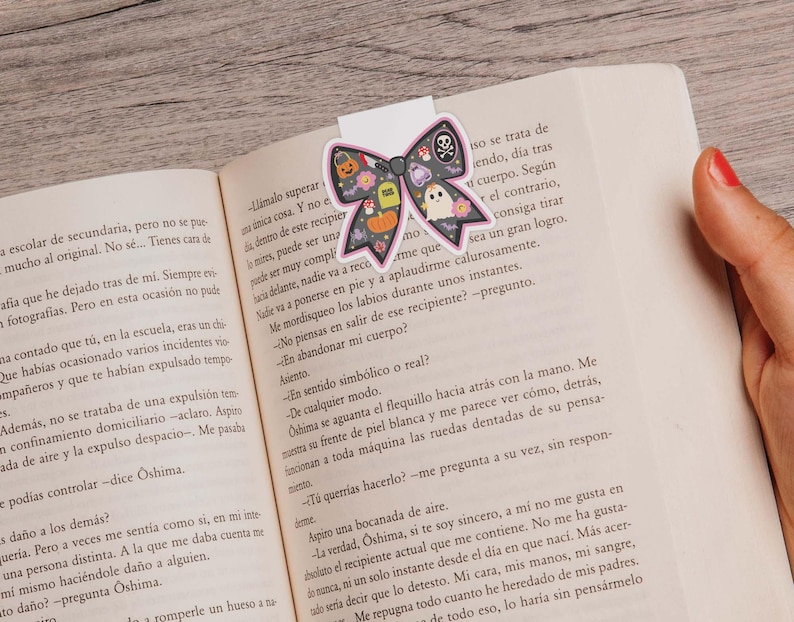 May include: A Halloween bow-shaped bookmark featuring a skull, ghost, pumpkin, and other icons. The bookmark is on an open book with text.