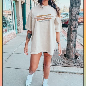 May include: A person wearing a light brown t-shirt with the text "Normalize Healing Your Childhood Trauma" in rainbow colors. The person is standing on a sidewalk in front of a tree.