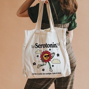 Może przedstawiać: Biała torba z pł&oacute;tna z kresk&oacute;wkową postacią kwiatu w okularach przeciwsłonecznych i napisem "Serotonin." Torba ma r&oacute;wnież napis "Go for a walk. Get sunlight. Take it slow."