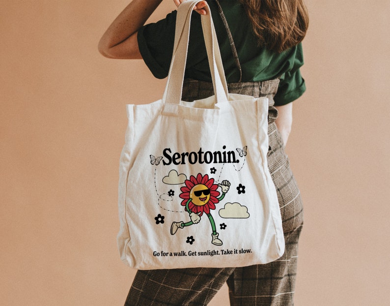 Może przedstawiać: Biała torba z pł&oacute;tna z kresk&oacute;wkową postacią kwiatu w okularach przeciwsłonecznych i napisem "Serotonin." Torba ma r&oacute;wnież napis "Go for a walk. Get sunlight. Take it slow."