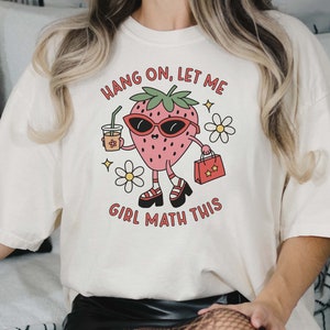 May include: White t-shirt with a cartoon strawberry wearing sunglasses, holding a cup of coffee and a shopping bag. The text on the shirt reads "Hang on, let me girl math this."