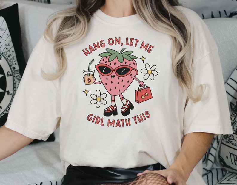May include: White t-shirt with a cartoon strawberry wearing sunglasses, holding a cup of coffee and a shopping bag. The text on the shirt reads "Hang on, let me girl math this."