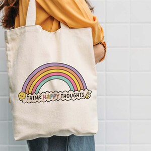 May include: A white canvas tote bag with a rainbow and the text "Think Happy Thoughts" in colorful letters. The rainbow has a yellow smiley face at the bottom left and a yellow star at the bottom right.