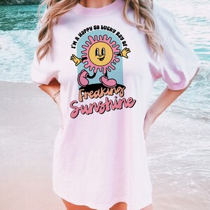 Op de afbeelding: Een roze t-shirt met een afbeelding van een lachende zon met de tekst "I'm a happy go lucky ray of freaking sunshine" erop gedrukt.