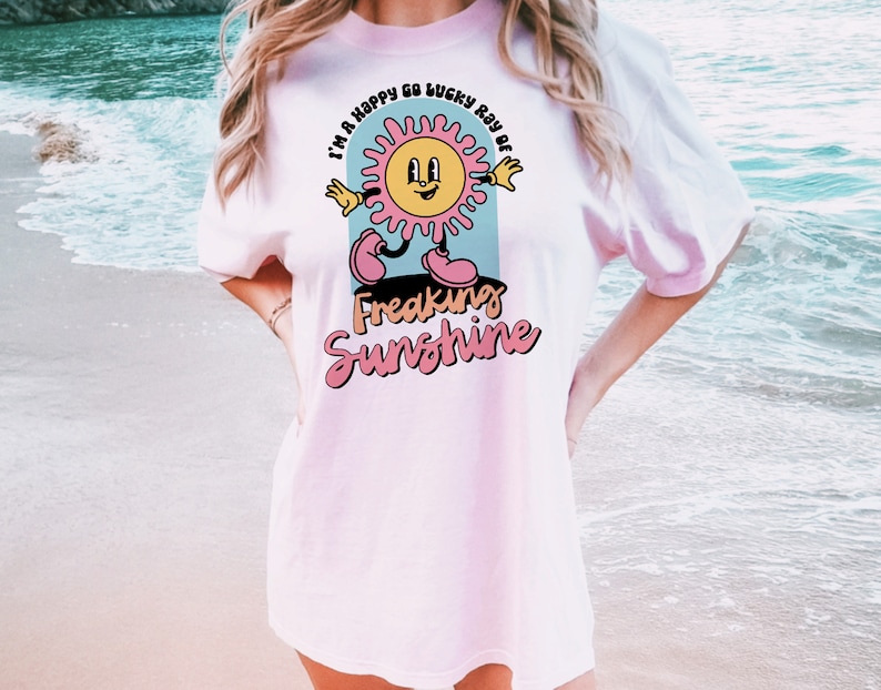 Op de afbeelding: Een roze t-shirt met een afbeelding van een lachende zon met de tekst "I'm a happy go lucky ray of freaking sunshine" erop gedrukt.
