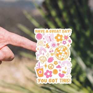 Puede incluir: Una pegatina blanca con un dise&ntilde;o rosa y amarillo. Presenta una variedad de ilustraciones que incluyen un arco&iacute;ris, un hongo, una fresa, un signo de paz, una corona, un rayo, una estrella, un coraz&oacute;n, una mariposa, una cara sonriente, una cereza, una taza de caf&eacute; y una bola de discoteca. El texto en la pegatina dice "Have a great day! You got this!"