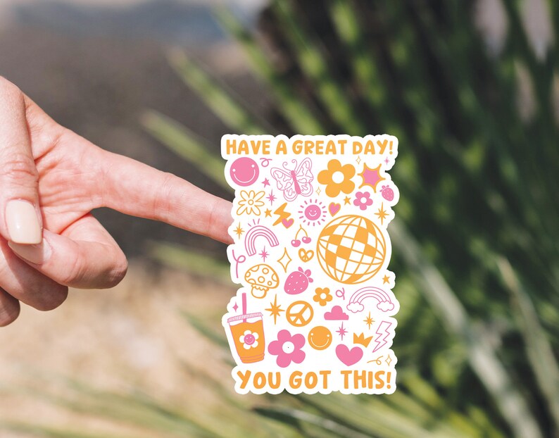 Puede incluir: Una pegatina blanca con un dise&ntilde;o rosa y amarillo. Presenta una variedad de ilustraciones que incluyen un arco&iacute;ris, un hongo, una fresa, un signo de paz, una corona, un rayo, una estrella, un coraz&oacute;n, una mariposa, una cara sonriente, una cereza, una taza de caf&eacute; y una bola de discoteca. El texto en la pegatina dice "Have a great day! You got this!"