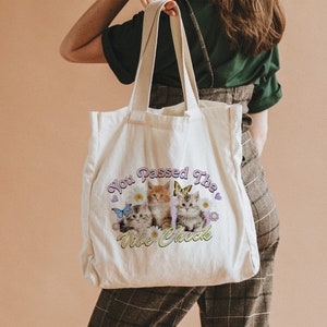Op de afbeelding: Een witte canvas tote bag met een afbeelding van drie kittens en vlinders. De tekst op de tas is "You Passed The Vibe Check".