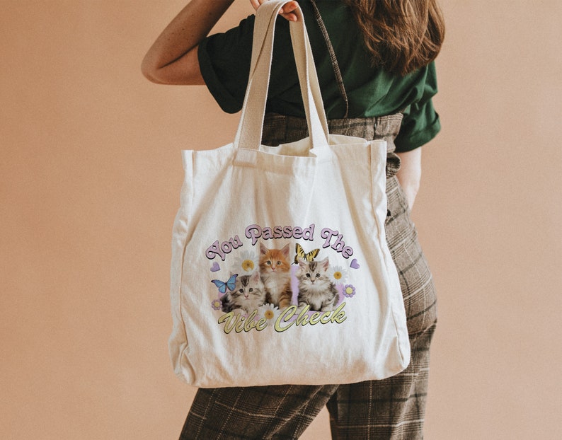 Op de afbeelding: Een witte canvas tote bag met een afbeelding van drie kittens en vlinders. De tekst op de tas is "You Passed The Vibe Check".