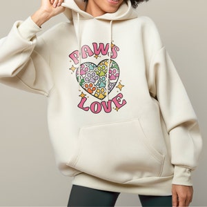 May include: A cream-colored hoodie with a pink heart design featuring paw prints in rainbow colors. The text "PAWS LOVE" is printed on the hoodie.