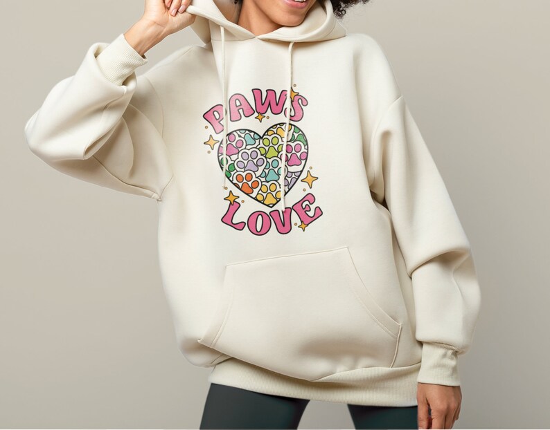 May include: A cream-colored hoodie with a pink heart design featuring paw prints in rainbow colors. The text "PAWS LOVE" is printed on the hoodie.