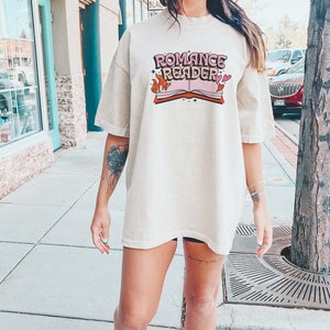 May include: A woman wearing a light beige oversized t-shirt with the text "Romance Reader" and a graphic of a book with flames and hearts.