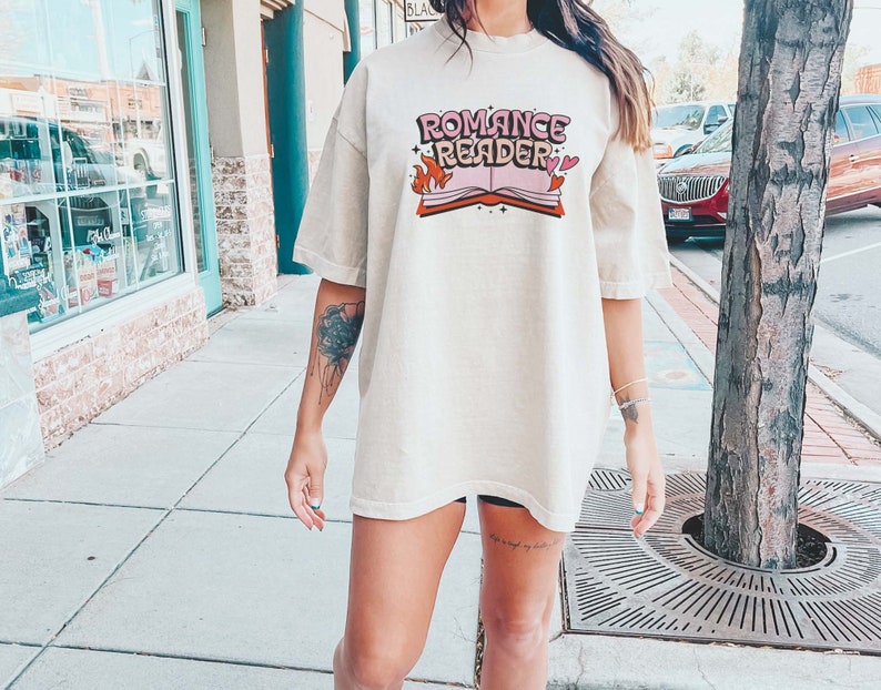 May include: A woman wearing a light beige oversized t-shirt with the text "Romance Reader" and a graphic of a book with flames and hearts.