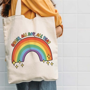 K&ouml;nnte beinhalten: Eine wei&szlig;e Canvas-Tragetasche mit einem Regenbogen und dem Text "Queer, All Day, All Year".