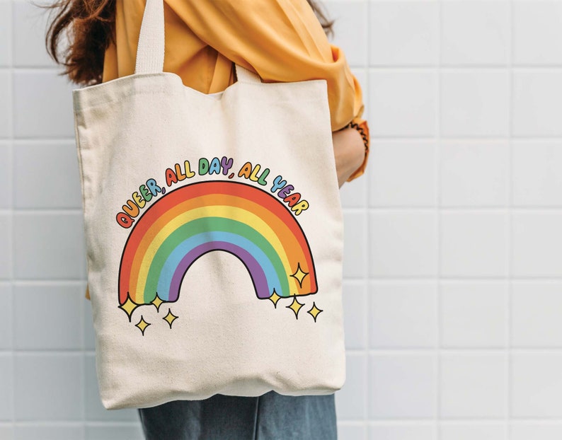 K&ouml;nnte beinhalten: Eine wei&szlig;e Canvas-Tragetasche mit einem Regenbogen und dem Text "Queer, All Day, All Year".