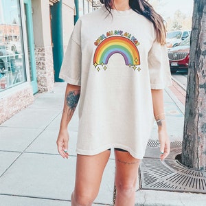 K&ouml;nnte beinhalten: Eine Frau tr&auml;gt ein hellbeiges T-Shirt mit einem Regenbogen-Motiv und dem Text "Queer All Day, All Year".