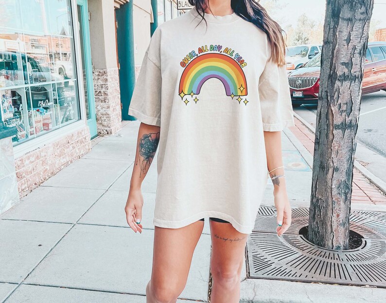 K&ouml;nnte beinhalten: Eine Frau tr&auml;gt ein hellbeiges T-Shirt mit einem Regenbogen-Motiv und dem Text "Queer All Day, All Year".