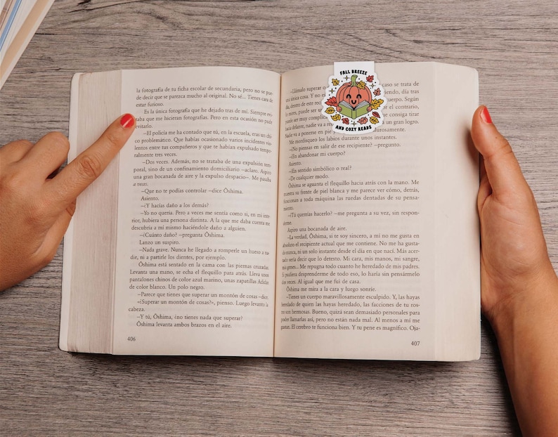 May include: A close-up of an open book with a bookmark featuring a pumpkin with a book and the text "Fall Bookish and Cozy Reads". The book is open to a page with text in Spanish.