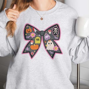 May include: A pale grey sweatshirt with a large black bow design, decorated with Halloween illustrations. The bow features a pumpkin, tombstone, ghost, and other spooky elements. The text "DEAD TIRED" is on the tombstone.