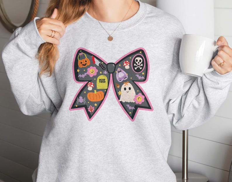 May include: A pale grey sweatshirt with a large black bow design, decorated with Halloween illustrations. The bow features a pumpkin, tombstone, ghost, and other spooky elements. The text "DEAD TIRED" is on the tombstone.
