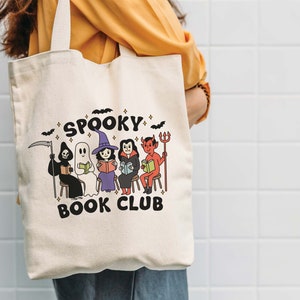 Puede incluir: Bolso tote de color crema con las palabras "Spooky Book Club" e ilustraciones de personajes de Halloween leyendo libros. Los personajes incluyen una parca, un fantasma, una bruja, un vampiro y un diablo. Murci&eacute;lagos y estrellas negras decoran la parte superior.