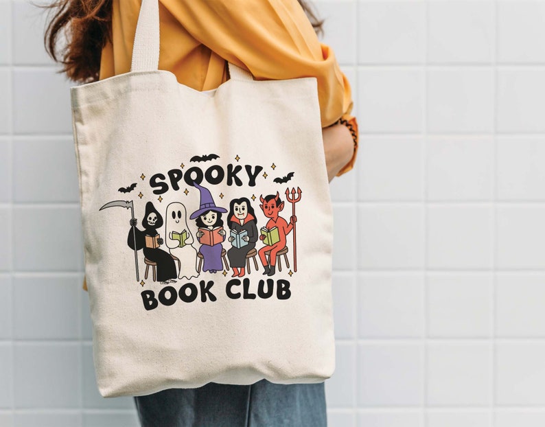 Puede incluir: Bolso tote de color crema con las palabras "Spooky Book Club" e ilustraciones de personajes de Halloween leyendo libros. Los personajes incluyen una parca, un fantasma, una bruja, un vampiro y un diablo. Murci&eacute;lagos y estrellas negras decoran la parte superior.