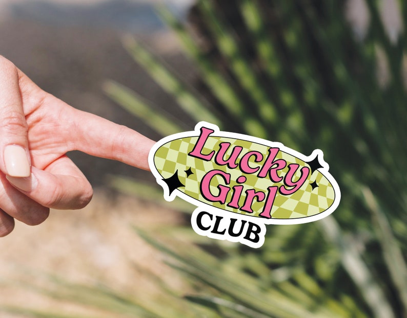 May include: A white sticker with a green and yellow checkered background. The text "Lucky Girl Club" is written in pink and yellow letters.