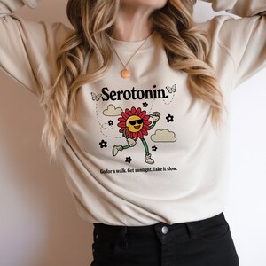 Może przedstawiać: Kremowa bluza z nadrukiem uśmiechniętego kwiatu w okularach przeciwsłonecznych i tekstem "Serotonin." Kwiat otoczony jest motylami i chmurami. Tekst pod nadrukiem brzmi "Go for a walk. Get sunlight. Take it slow."