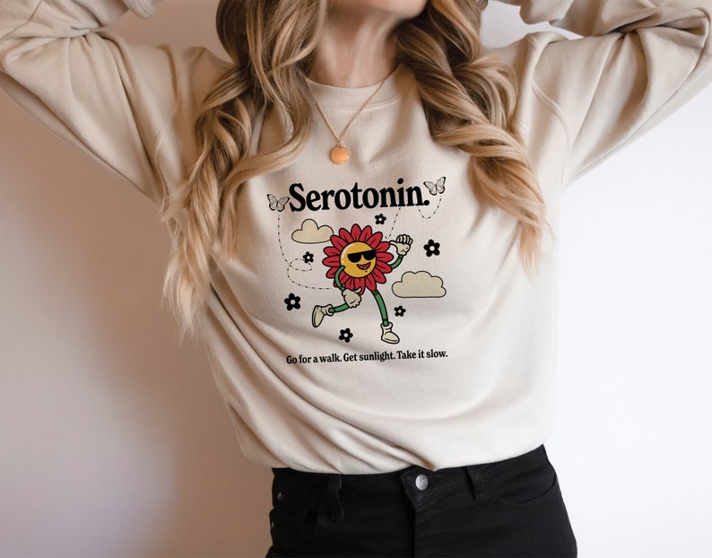 Może przedstawiać: Kremowa bluza z nadrukiem uśmiechniętego kwiatu w okularach przeciwsłonecznych i tekstem "Serotonin." Kwiat otoczony jest motylami i chmurami. Tekst pod nadrukiem brzmi "Go for a walk. Get sunlight. Take it slow."