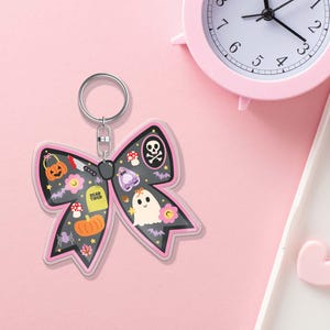 May include: A Halloween-themed bow-shaped keyring with a black background and pink trim. The keyring features various spooky illustrations, including a jack-o'-lantern, ghost, and skull. The words "Dead Tired" are also visible.