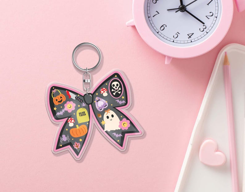 May include: A Halloween-themed bow-shaped keyring with a black background and pink trim. The keyring features various spooky illustrations, including a jack-o'-lantern, ghost, and skull. The words "Dead Tired" are also visible.