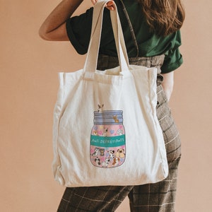 Pu&ograve; includere: Una borsa tote in tela bianca con un'illustrazione colorata di un barattolo pieno di animali dei cartoni animati e il testo "Anti-Depressants".