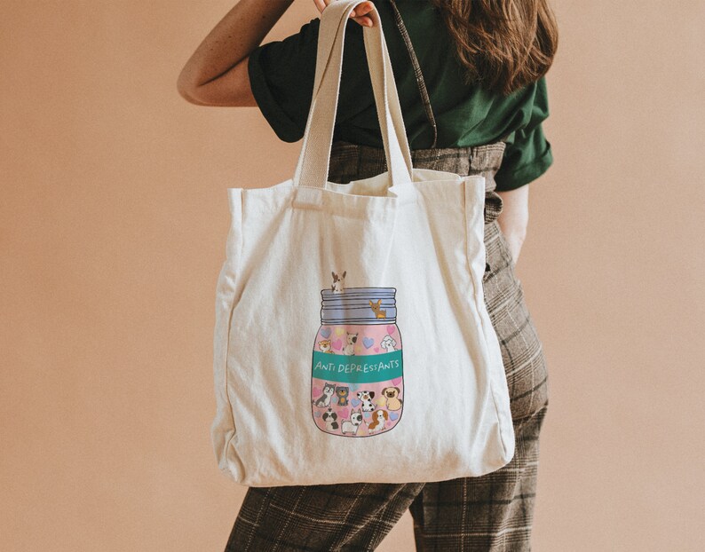 Pu&ograve; includere: Una borsa tote in tela bianca con un'illustrazione colorata di un barattolo pieno di animali dei cartoni animati e il testo "Anti-Depressants".