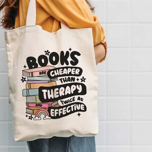 May include: A white canvas tote bag with a black and white graphic that says "Books are cheaper than therapy and twice as effective". The graphic also includes a stack of colorful books with a rainbow and stars.