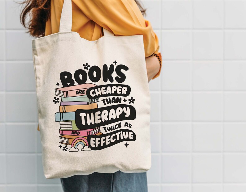 May include: A white canvas tote bag with a black and white graphic that says "Books are cheaper than therapy and twice as effective". The graphic also includes a stack of colorful books with a rainbow and stars.