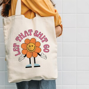 May include: A white canvas tote bag with a pink and orange flower graphic and the text "Let That Shit Go".