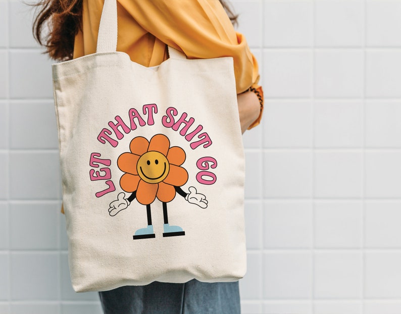May include: A white canvas tote bag with a pink and orange flower graphic and the text "Let That Shit Go".