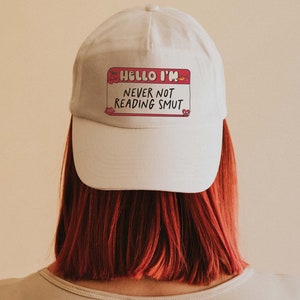 May include: White baseball cap with a pink name tag that reads "Hello I'm Never Not Reading Smut".