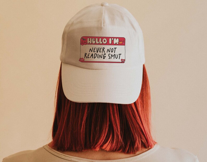 May include: White baseball cap with a pink name tag that reads "Hello I'm Never Not Reading Smut".